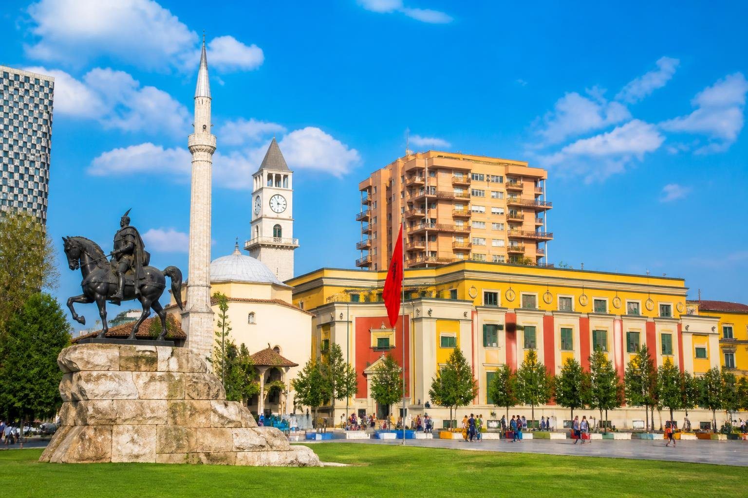 Vista panorâmica da cidade de Tirana com a praça Skanderbeg, edifícios coloridos, árvores e pessoas caminhando nas ruas.